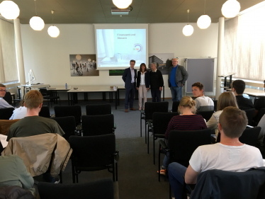 4 Personen vor Leinwand gucken zu mehreren Personen auf Stühlen